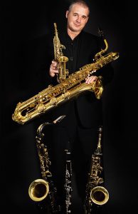 Andy Firth holding soprano saxophone with a clarinet and multiple saxophones on a stand.